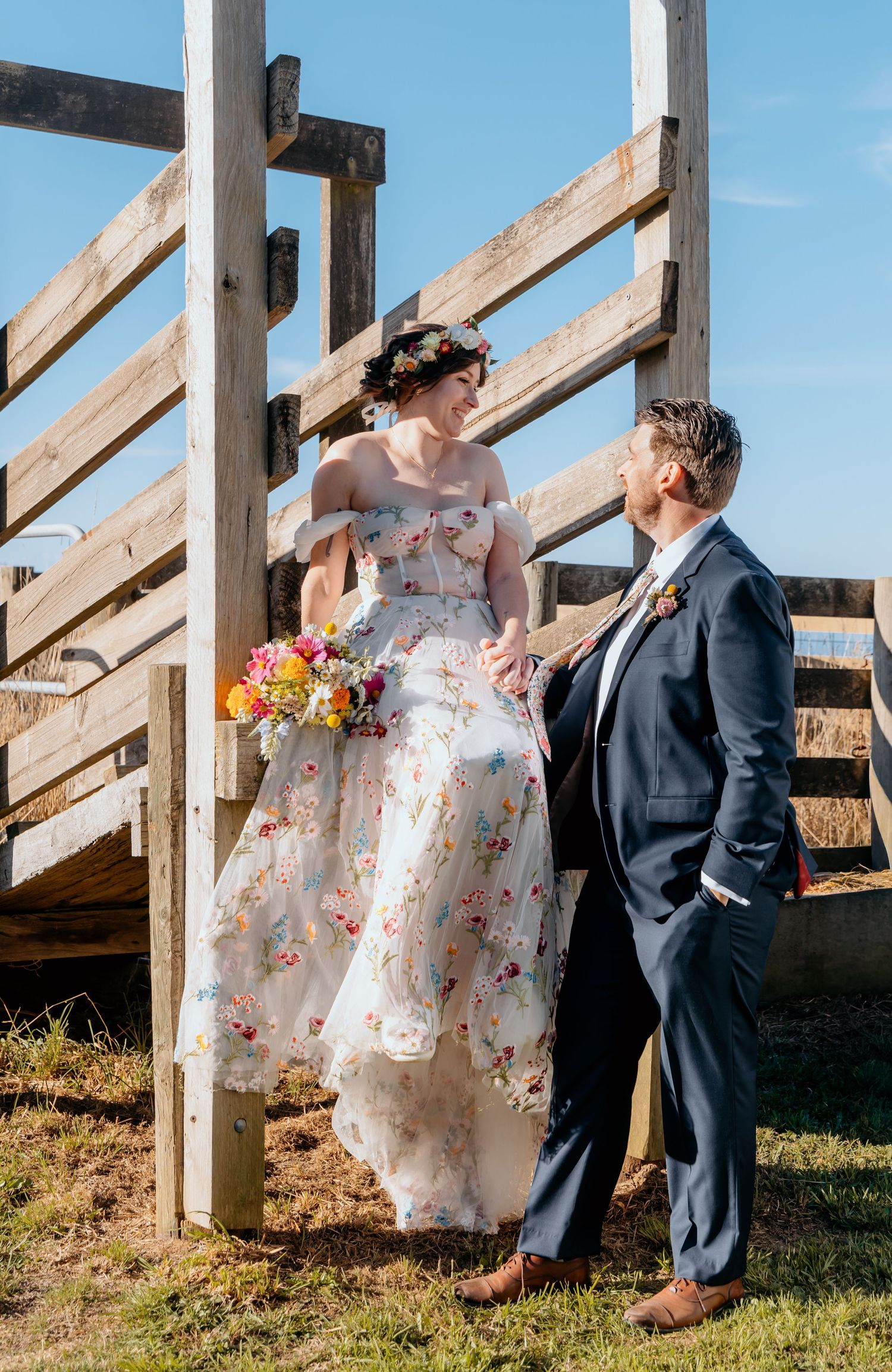 Bride in floral gown with flower crown holds bouquet while groom in navy suit looks up at her by wooden fence.