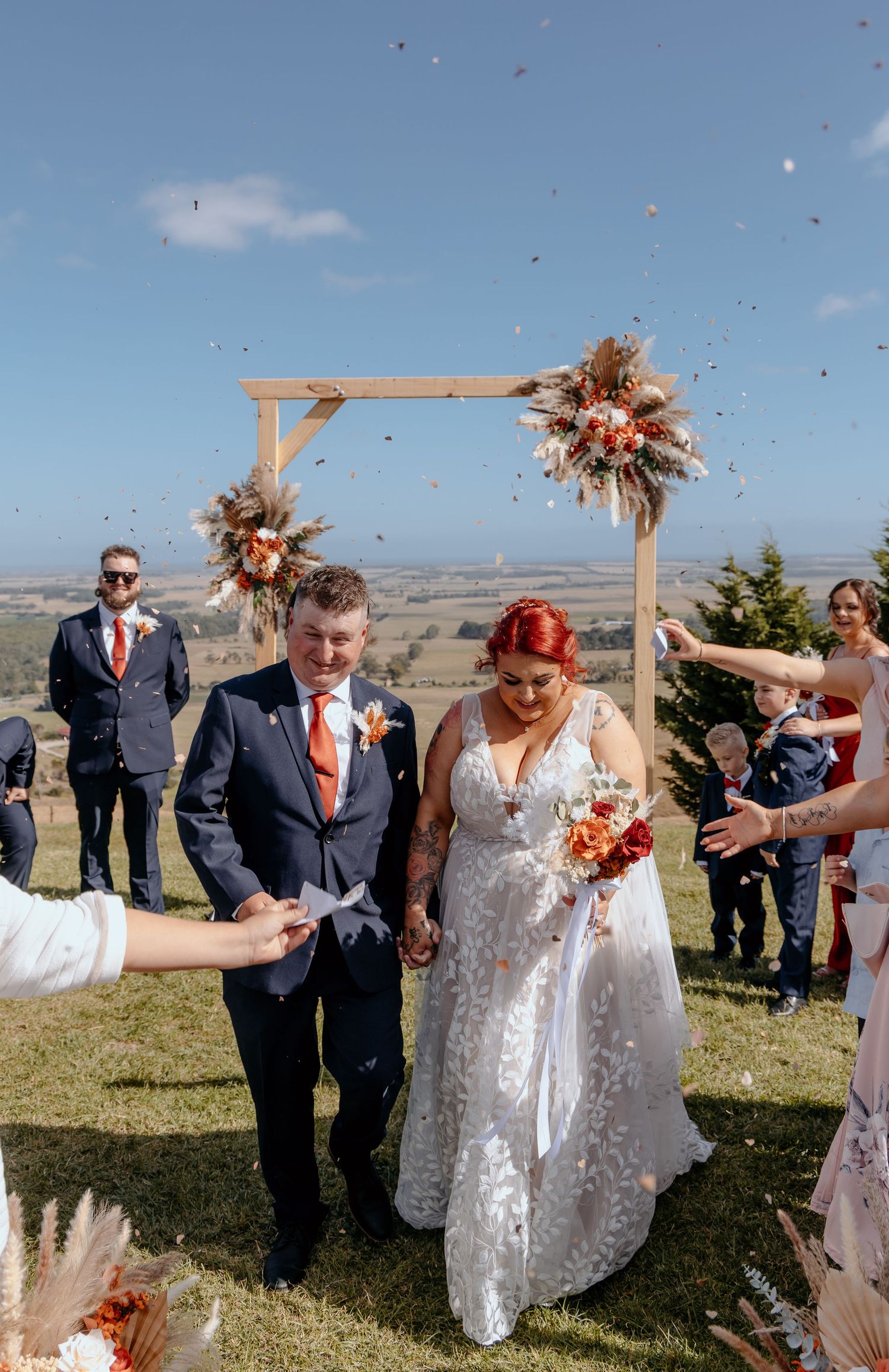 Bride and groom walk under wooden arch as confetti falls at outdoor hilltop wedding ceremony with scenic views.