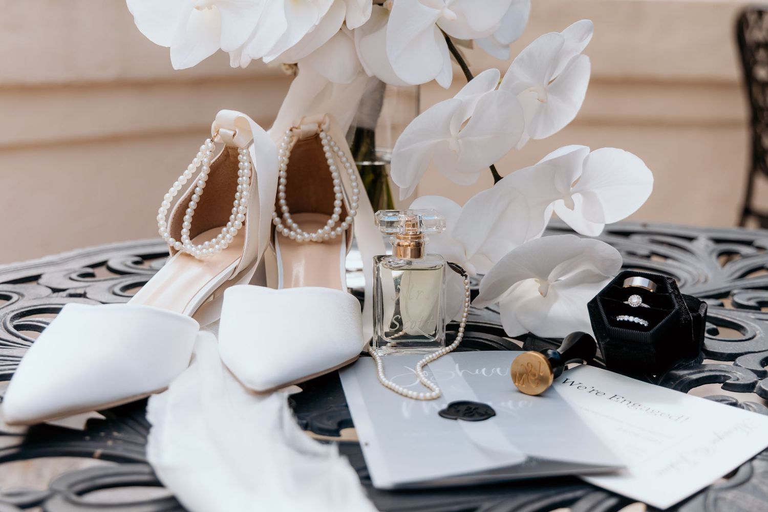 Elegant bridal flat lay featuring pearl heels, white pumps, perfume, ring box, and floral accents on ornate black table.