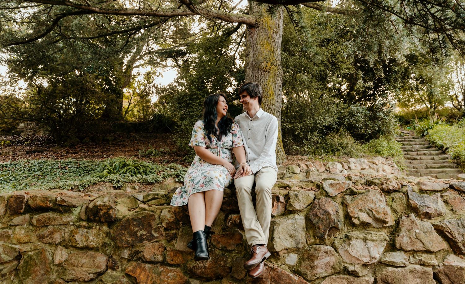 A couple sits intimately on a stone wall during an outdoor photoshoot surrounded by greenery.