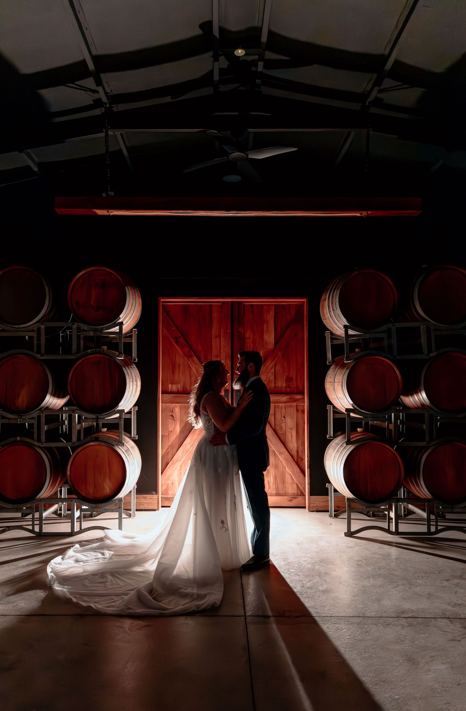 A silhouetted couple in wedding attire stand in a winery barrel room with dramatic red lighting and wooden barrels.