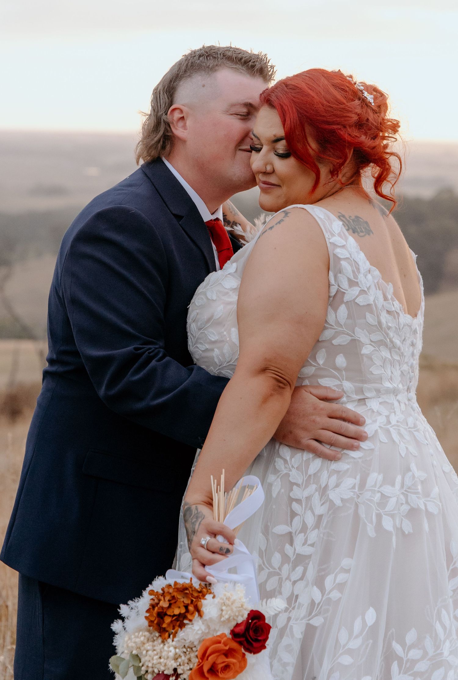 Groom in navy suit kisses bride with red hair wearing lace wedding dress holding autumn bouquet.