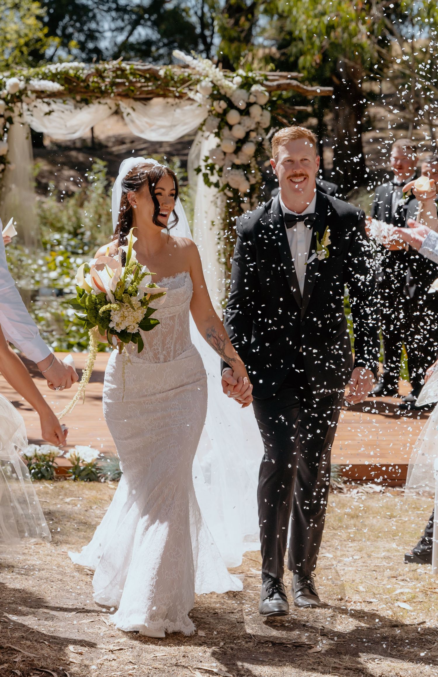Bride and groom walk hand-in-hand through a confetti shower after their outdoor wedding ceremony.