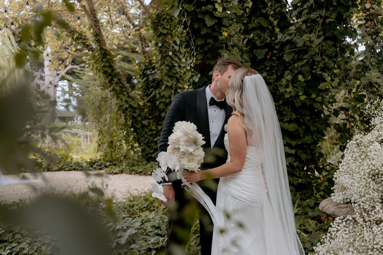 Bride and groom share a romantic kiss surrounded by lush green ivy walls, bride holding white floral bouquet.