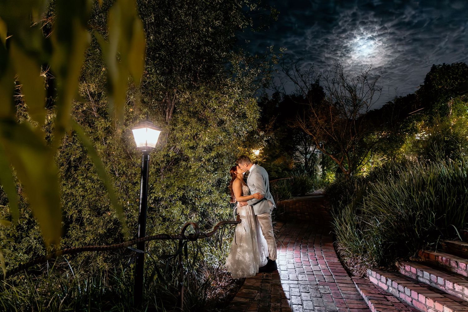 A couple stands on a brick path under a vintage streetlamp and full moon in a romantic garden at night.