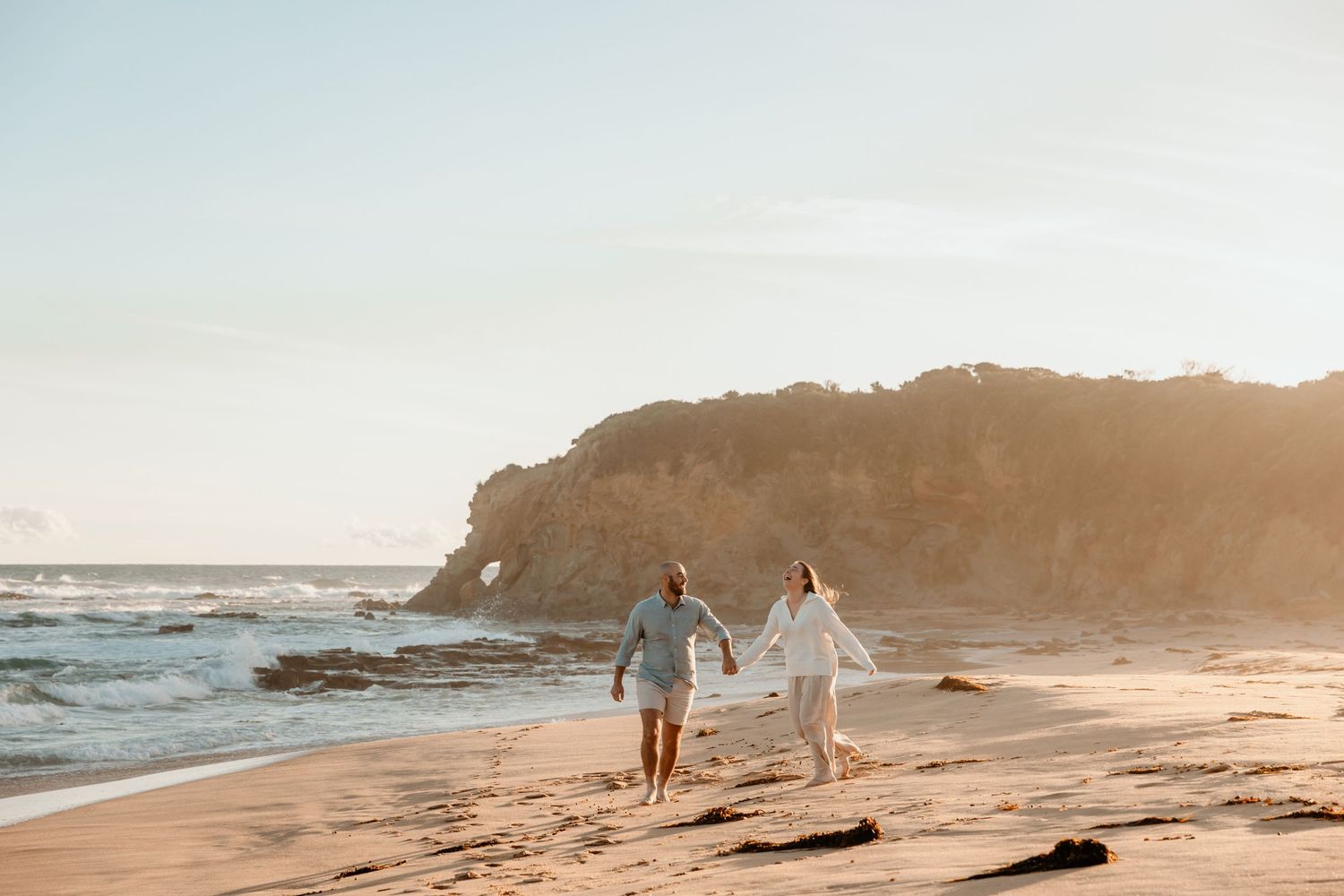 Couple walking hand in hand on a scenic beach with cliffs at sunset.
