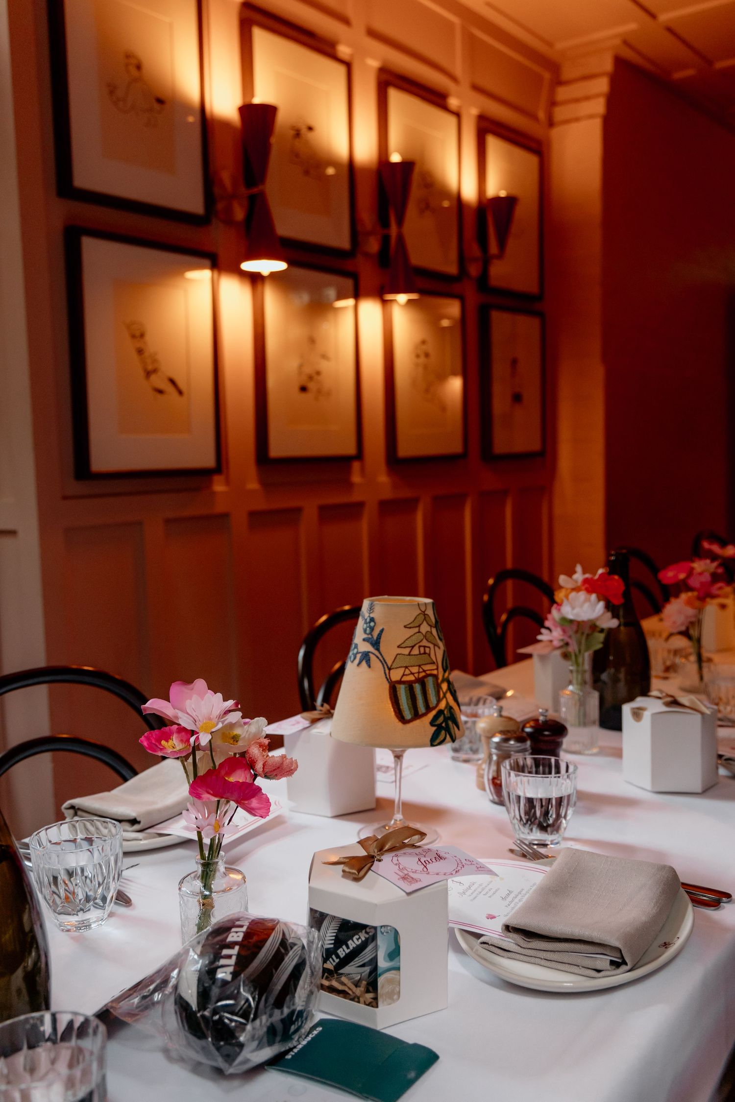 Elegant restaurant table setting with pink flowers, white tablecloth, and framed artwork on warm-lit wooden walls.