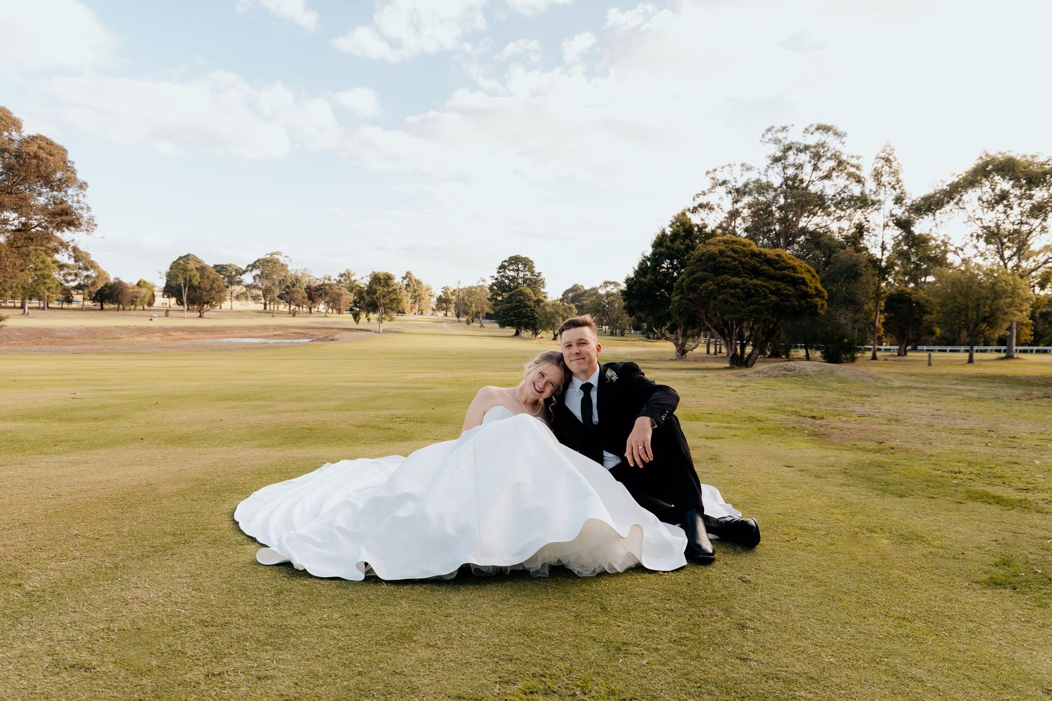 A newlywed couple sits together on a grassy golf course with trees in the background on their wedding day.