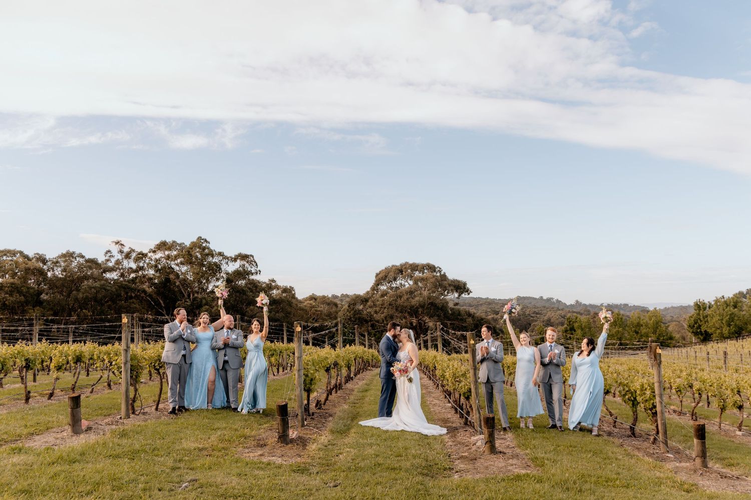 Wedding party in light blue dresses poses together in a scenic vineyard on a sunny afternoon.