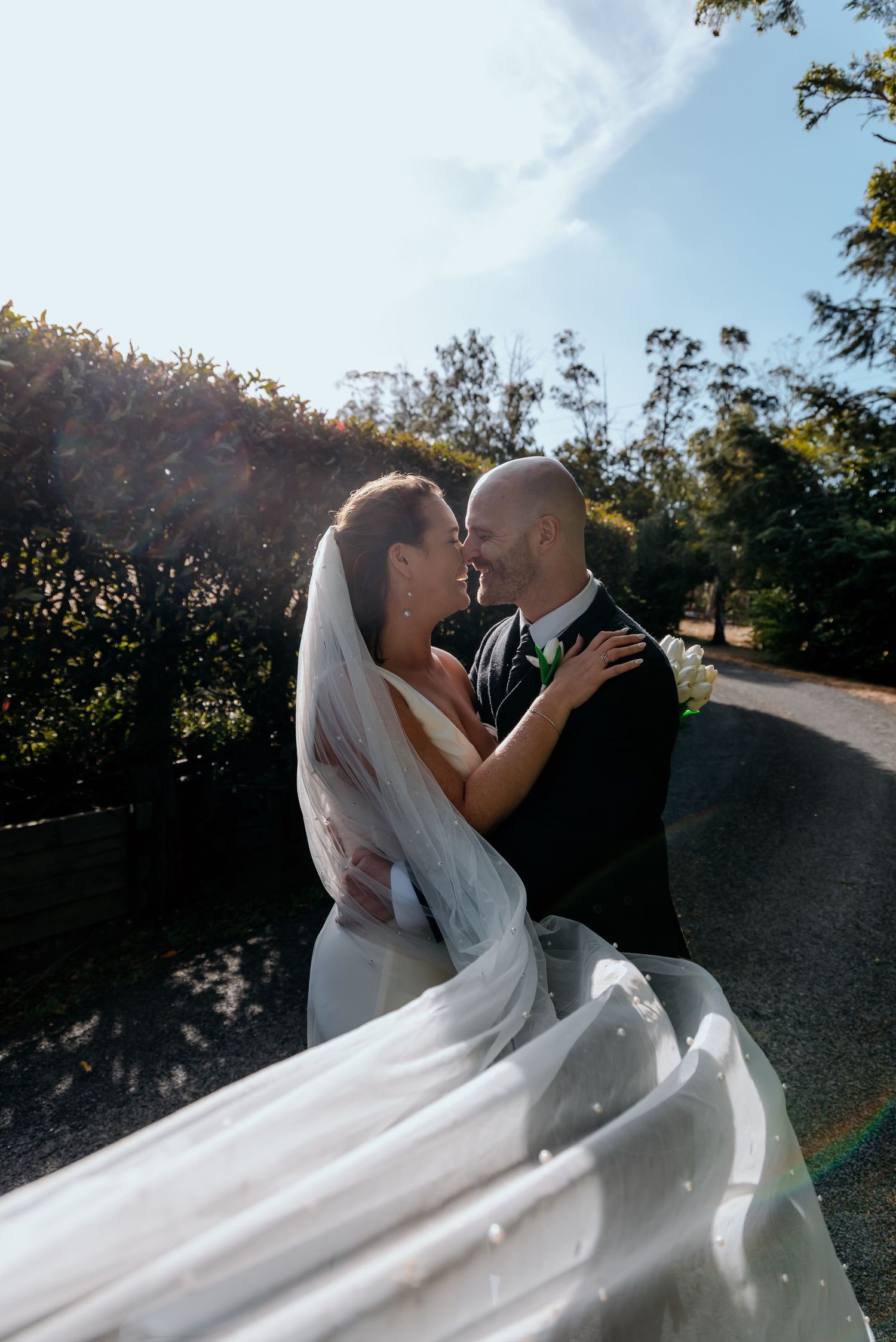 Bride and groom share a romantic moment outdoors, her veil flowing in the breeze on their wedding day.