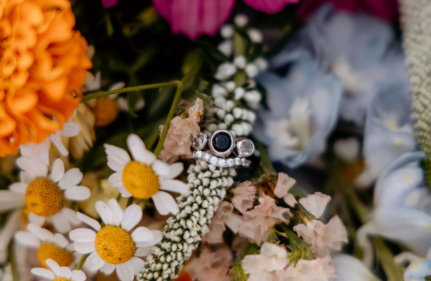 Elegant sapphire and diamond engagement rings resting among colorful wedding bouquet flowers including daisies and baby's breath.