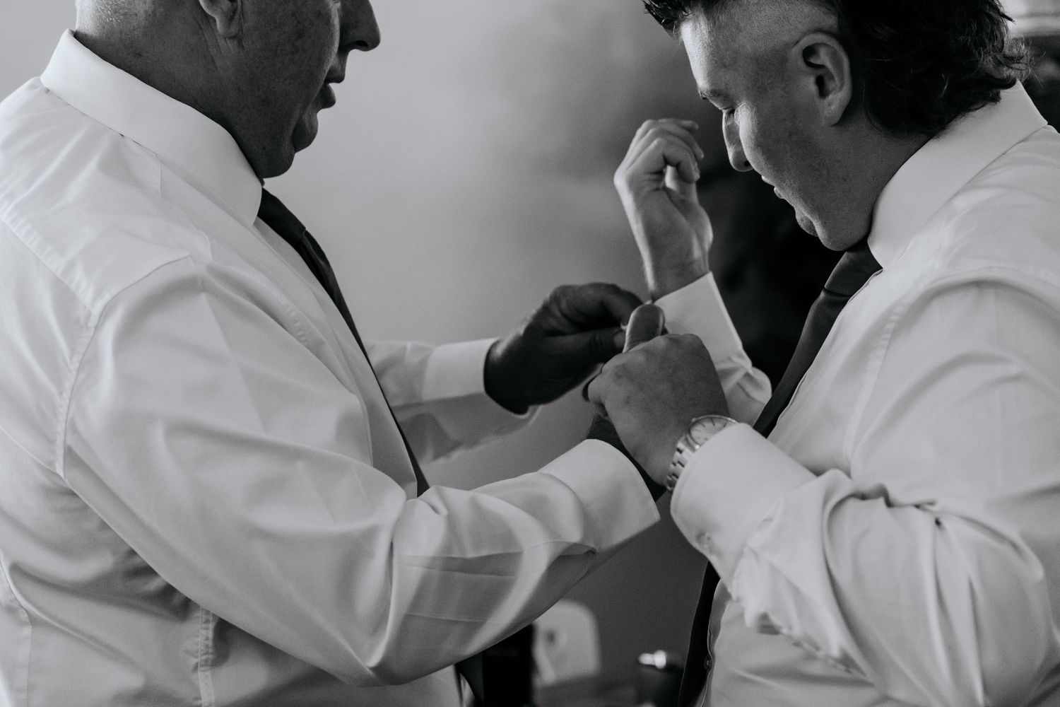 Two men in white dress shirts adjusting cufflinks and tie in black and white wedding preparation photo.