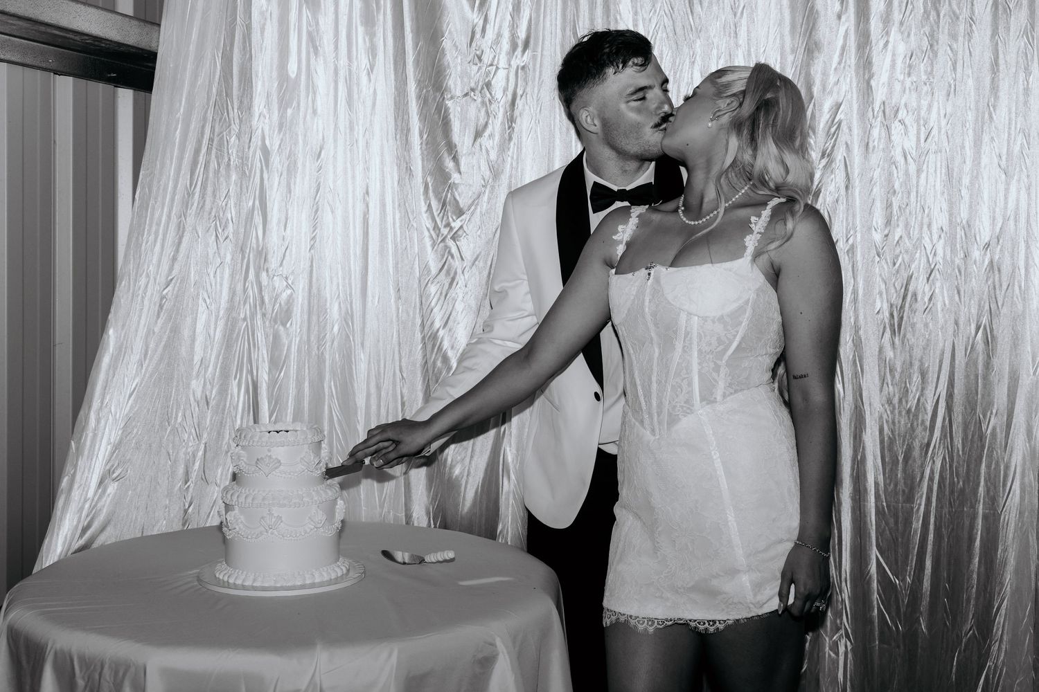 Black and white photo of a couple kissing while cutting their wedding cake in front of elegant draped curtains.