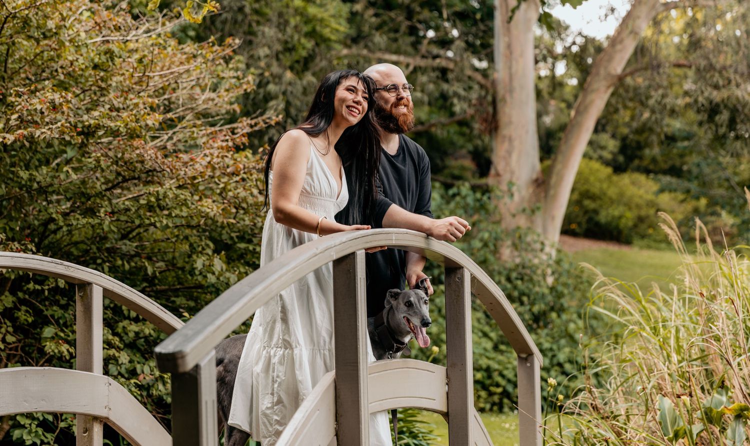 Couple in wedding attire standing on white garden bridge surrounded by autumn foliage.