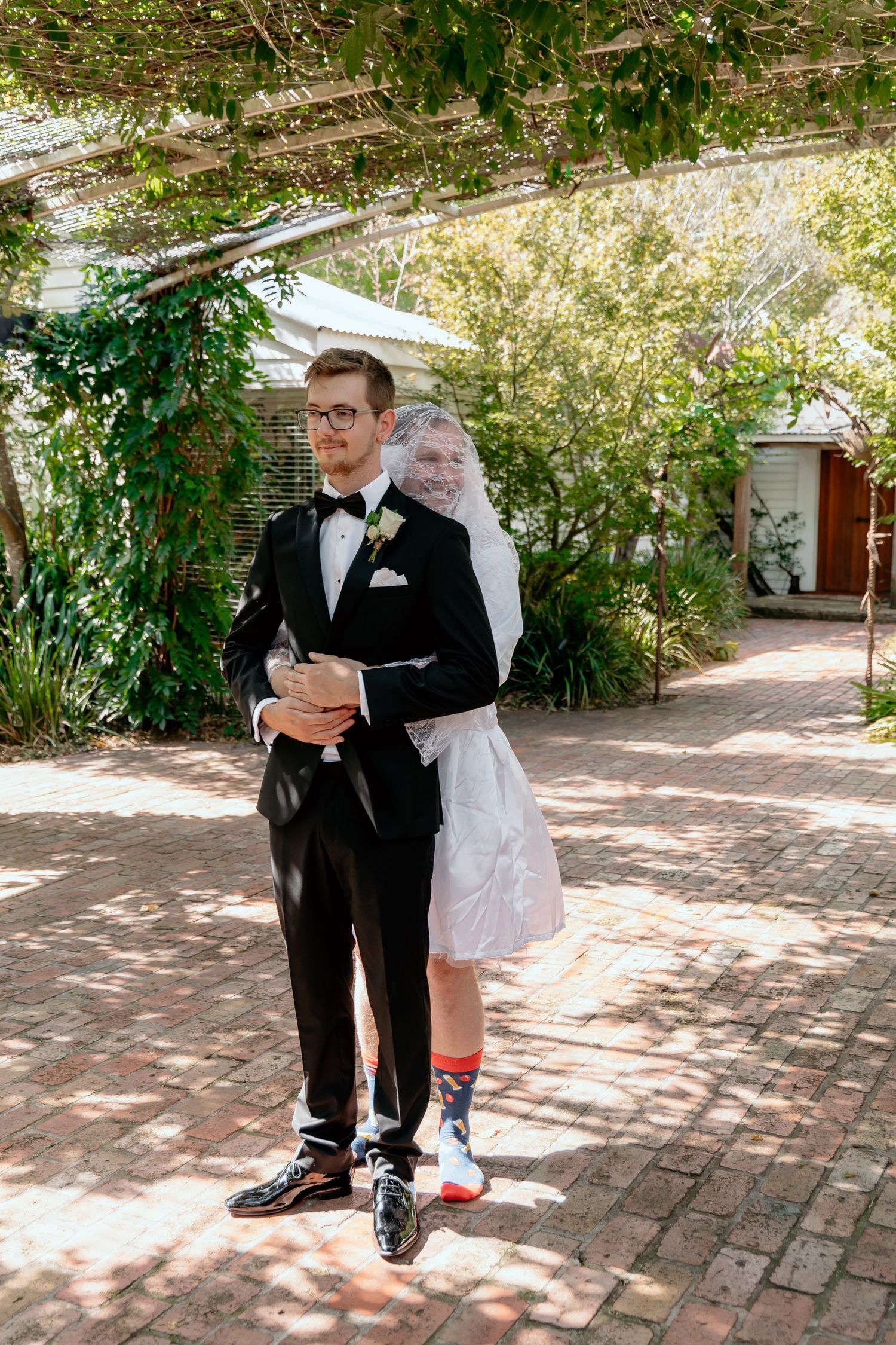 A couple in formal attire prepare for their wedding day in an outdoor garden setting with brick pathways.