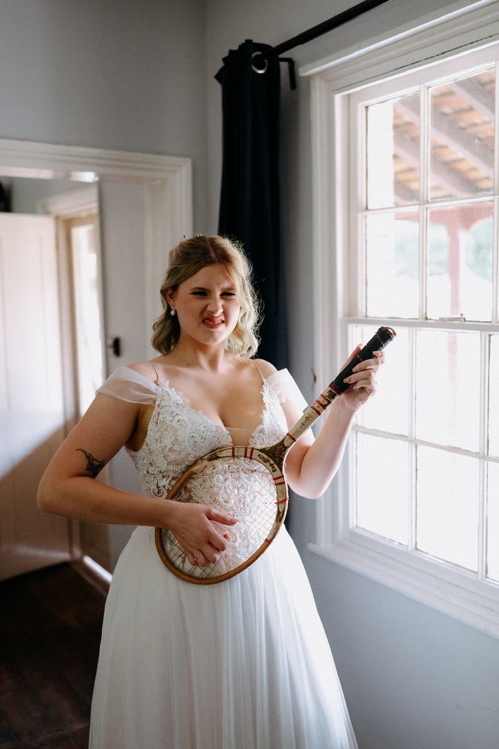 Bride in white dress holding banjo near window in bright indoor setting.