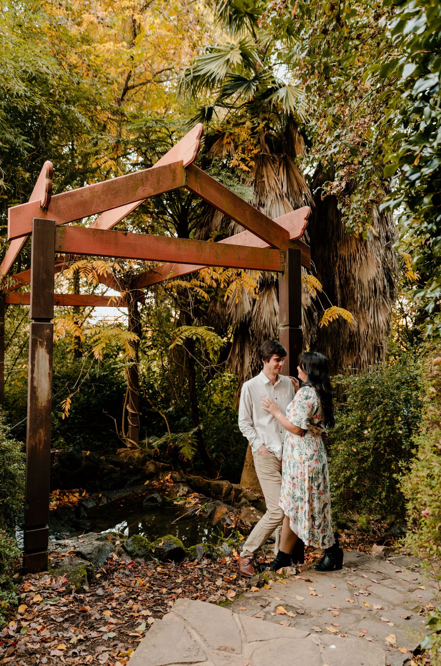 A romantic couple embraces under a wooden pergola surrounded by autumn foliage.