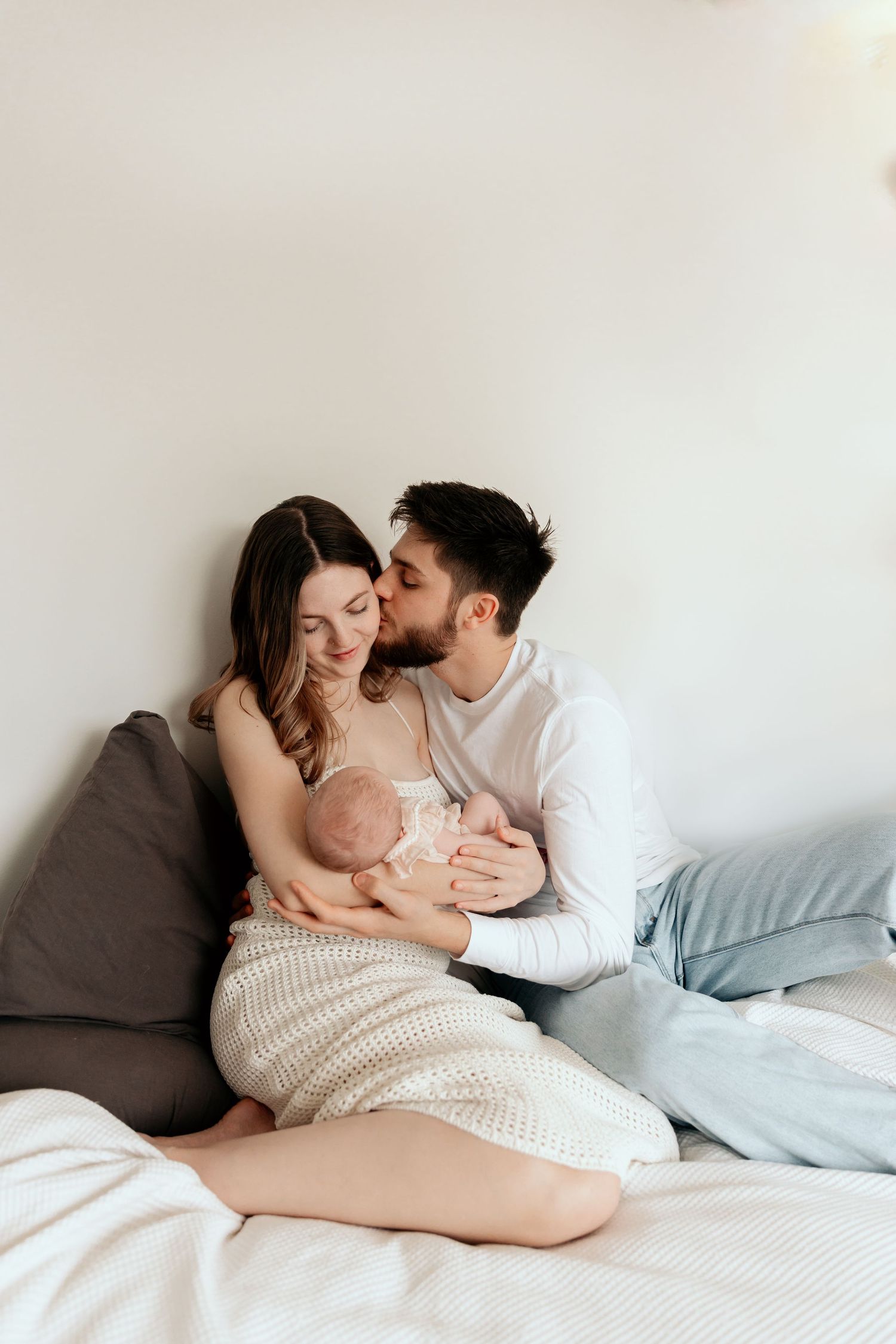 Tender family moment on cozy bed with baby in white knit dress.