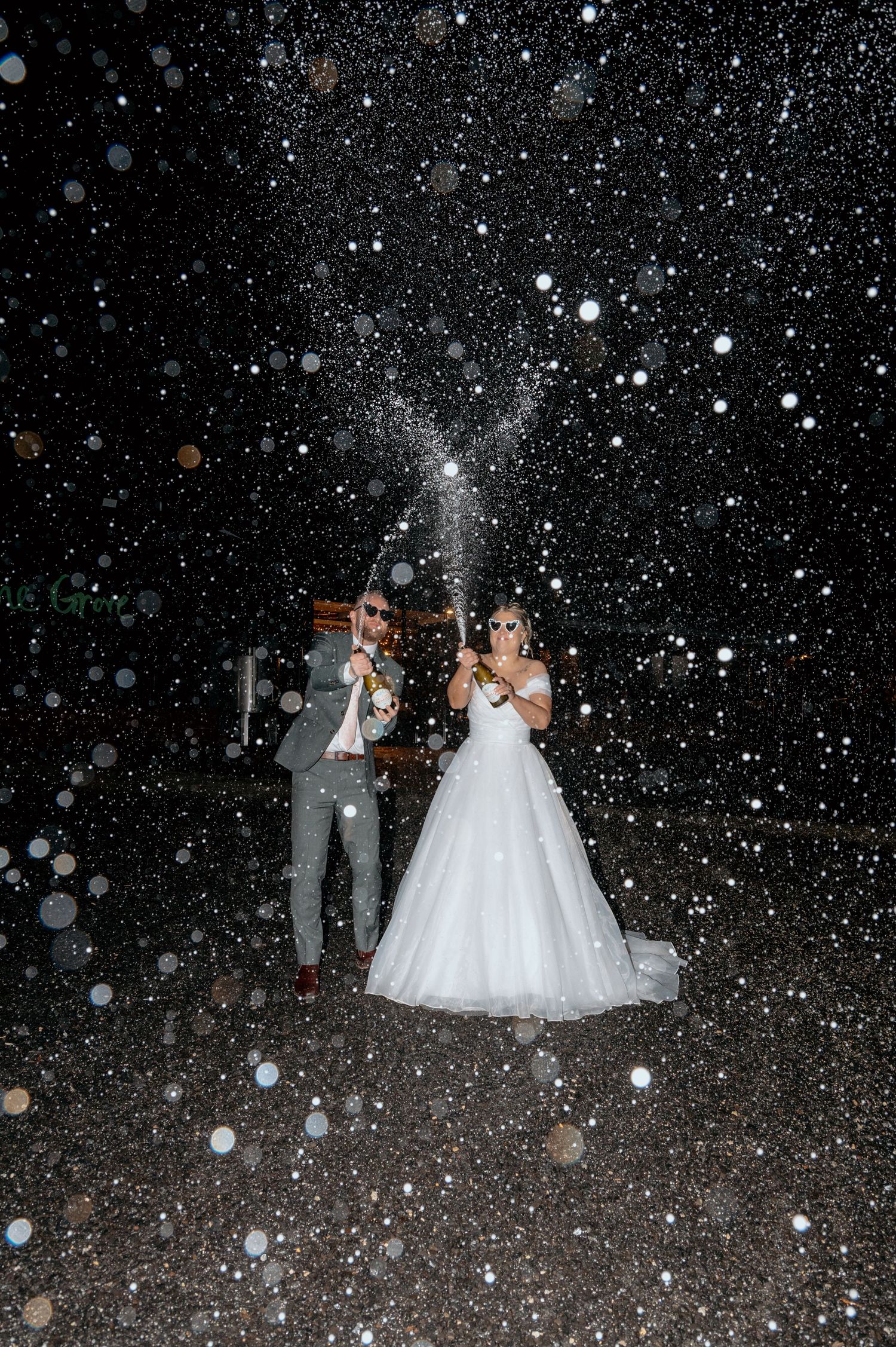 Bride and groom celebrating at night, popping champagne with sparkling droplets flying around them outdoors.