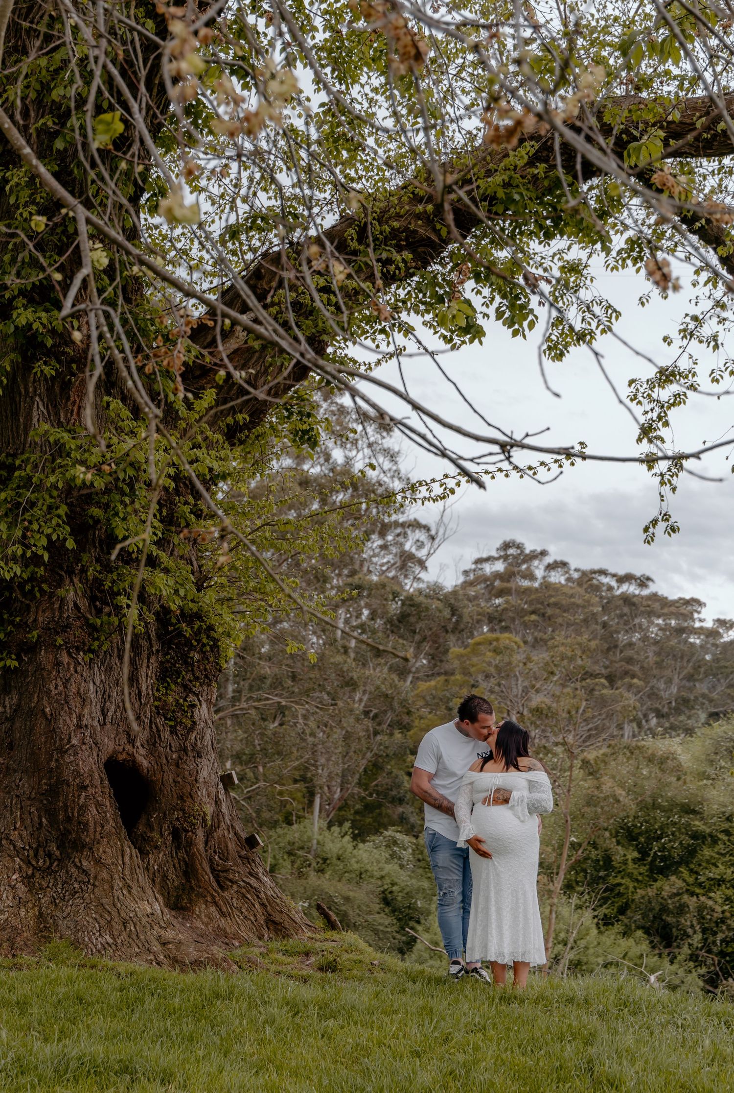 A couple walks together across a grassy hillside beneath towering trees on a cloudy day.