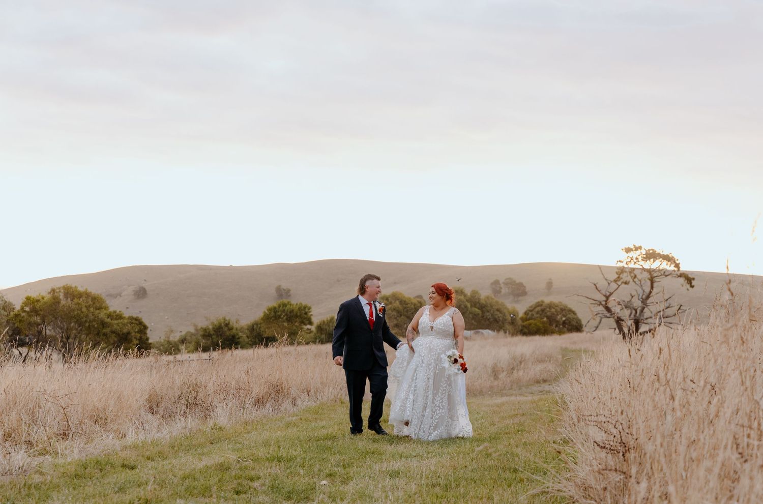 Bride and groom walk hand in hand through golden fields with rolling hills backdrop at outdoor wedding.