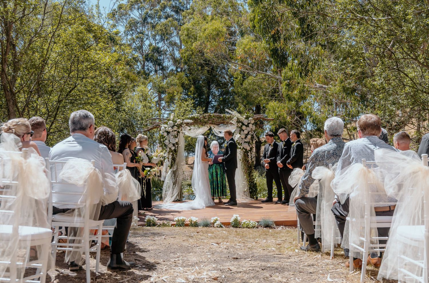 Outdoor wedding ceremony with guests seated in white chairs as couple exchanges vows beneath floral arch.