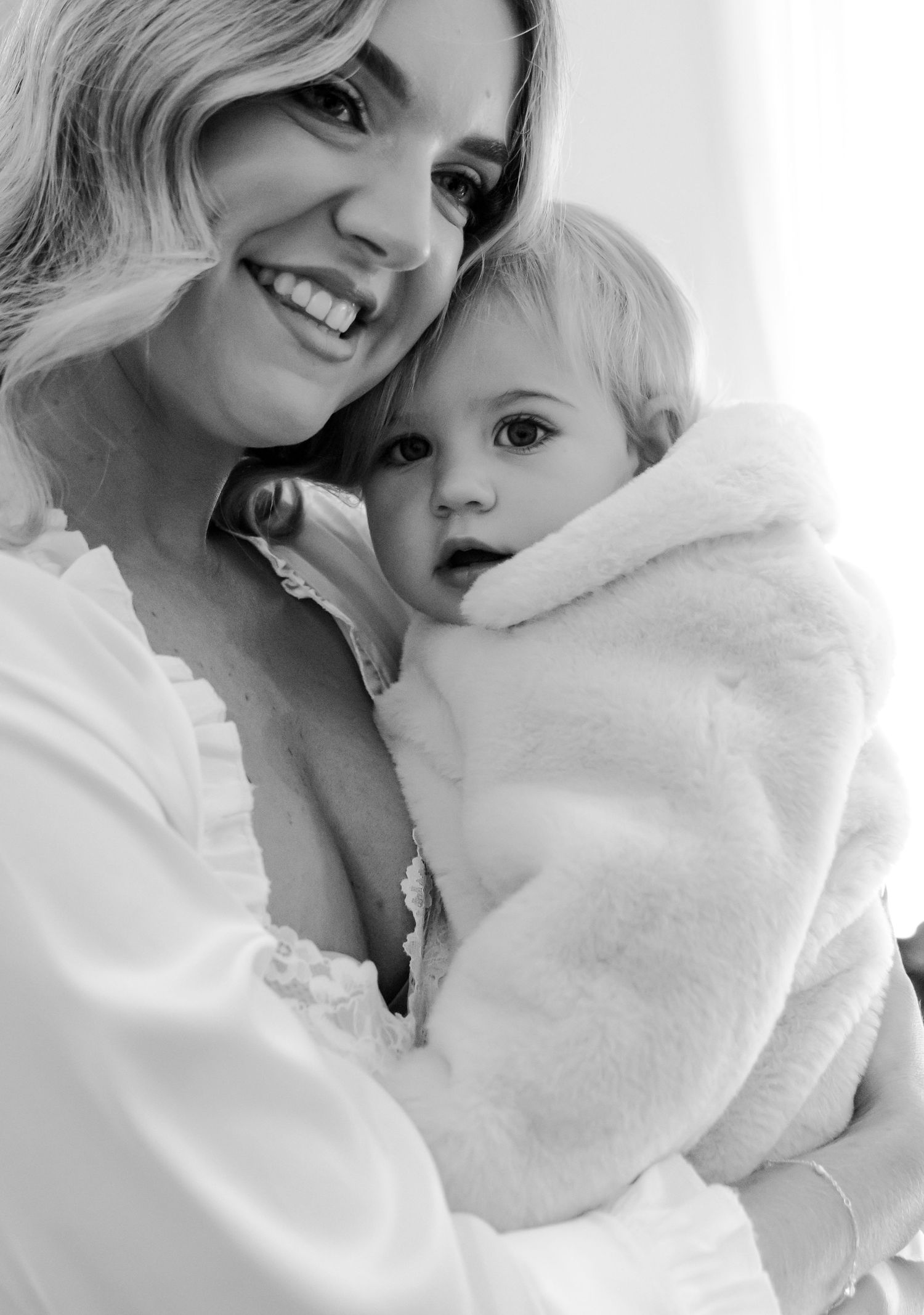 Black and white photo of a smiling woman holding a baby dressed in a fluffy white coat.