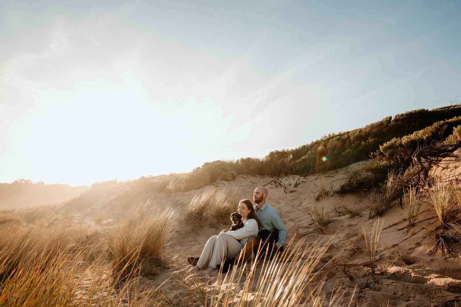 Couple sits in golden beach grass during sunset creating a dreamy coastal atmosphere.