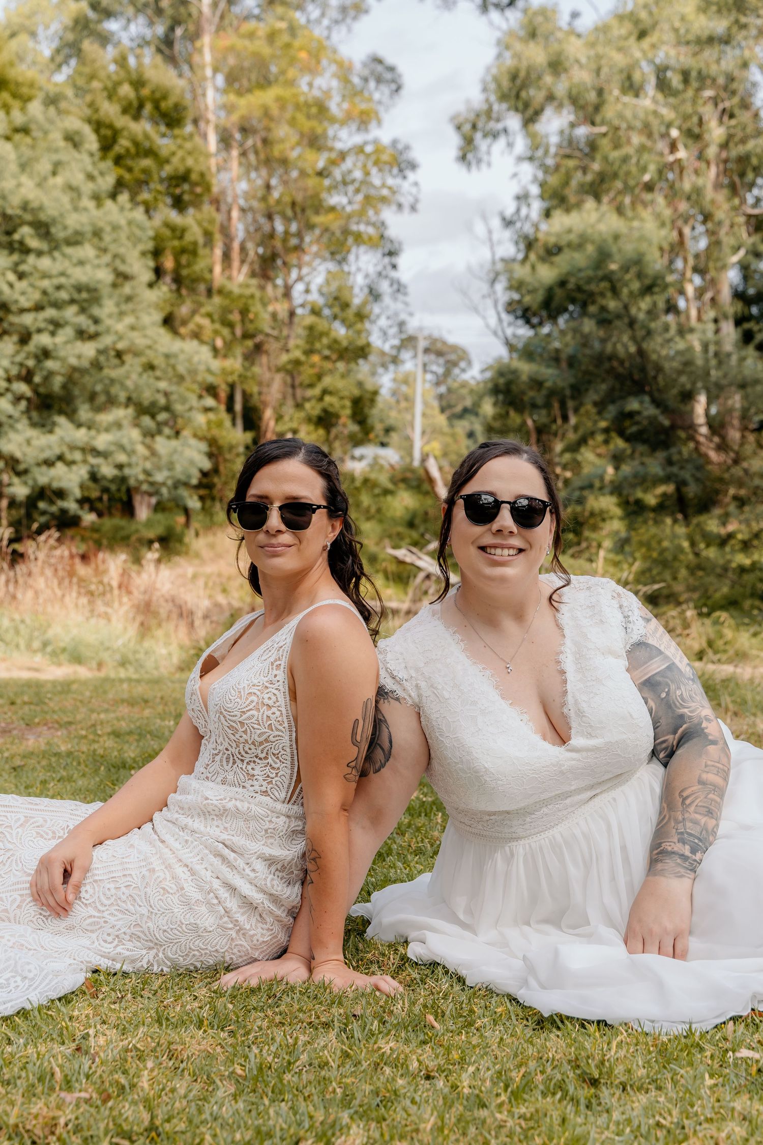 Two people in white dresses and sunglasses sit together on grass with eucalyptus trees in the background.