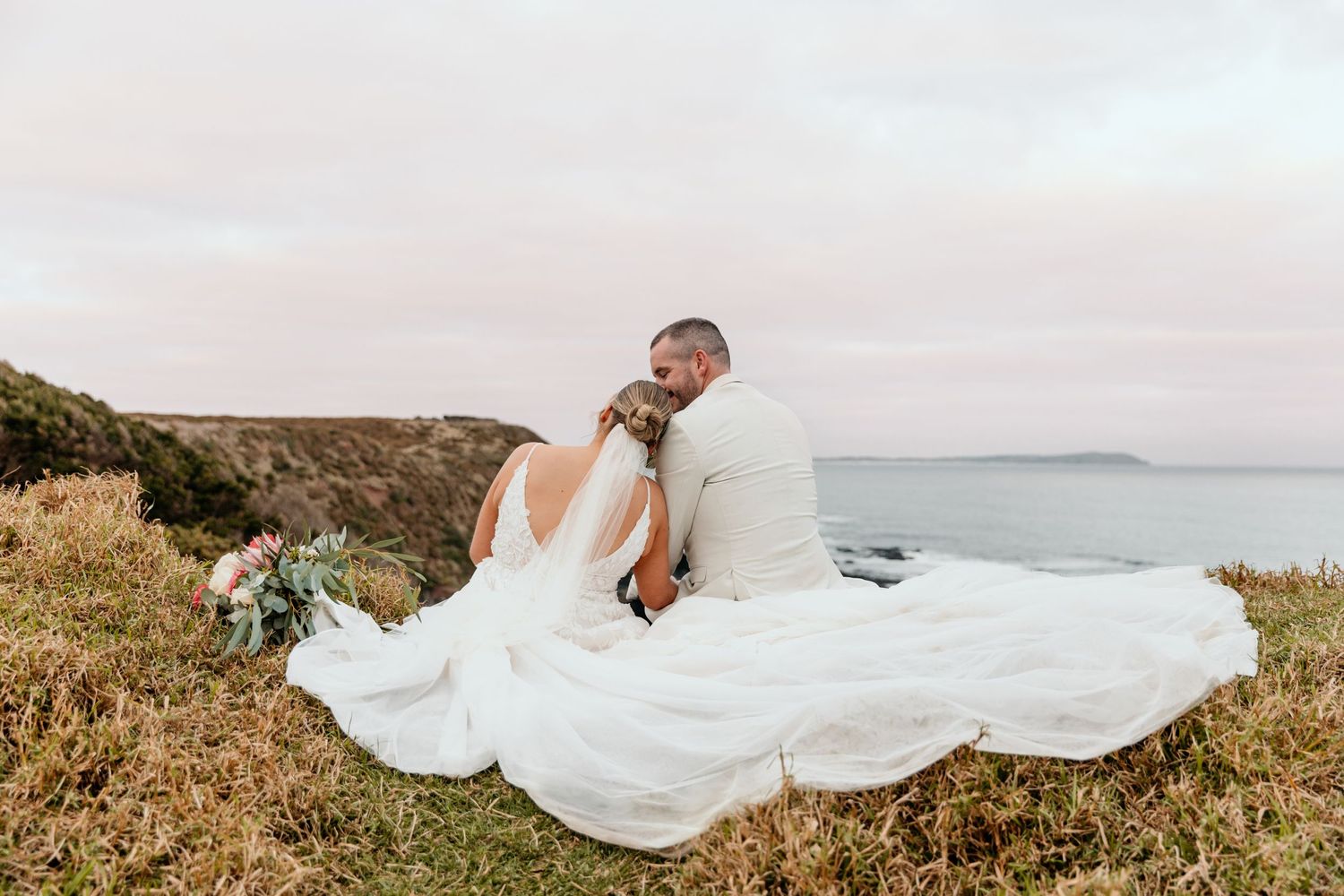 Couple sits on clifftop with flowing white dress billowing in the ocean breeze.