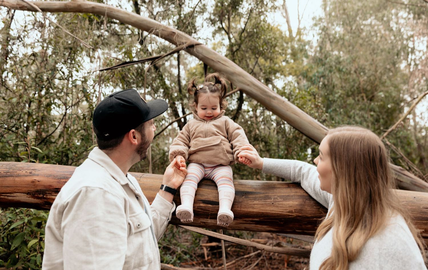A toddler sits on a fallen log while two adults interact playfully in a wooded setting during an outdoor photoshoot.