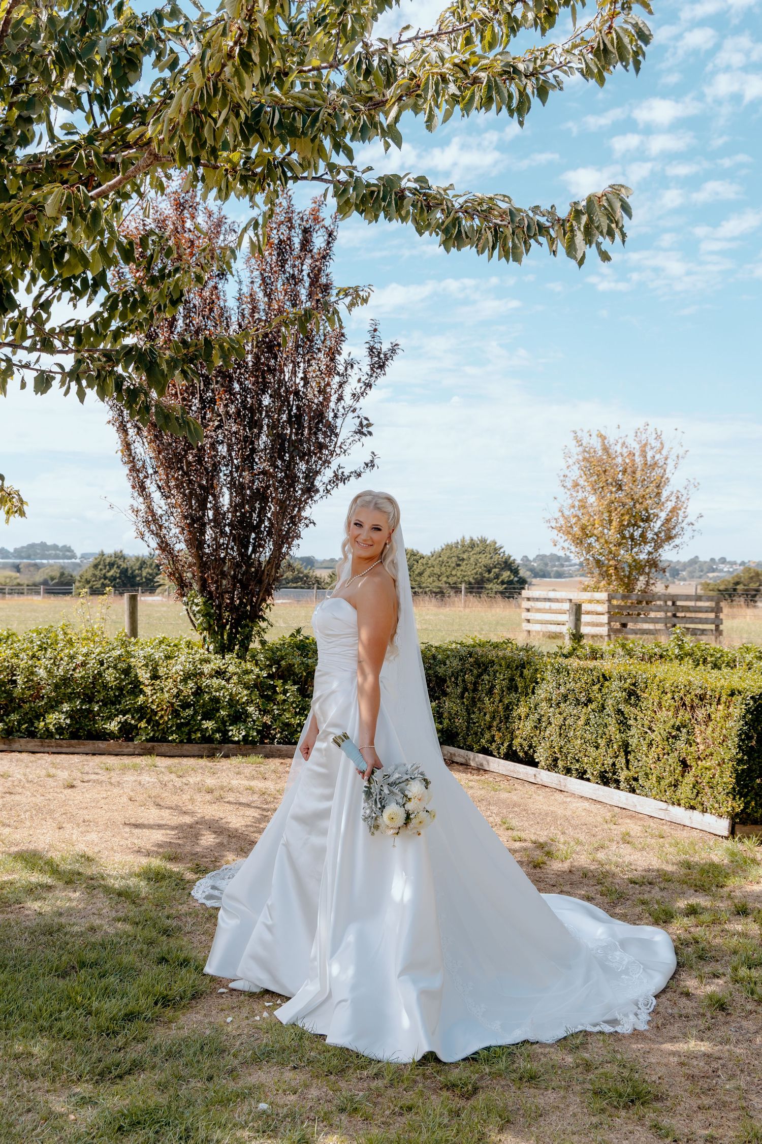 Blonde bride in a strapless white gown with long train holds bouquet outdoors on a sunny day at a farm venue.