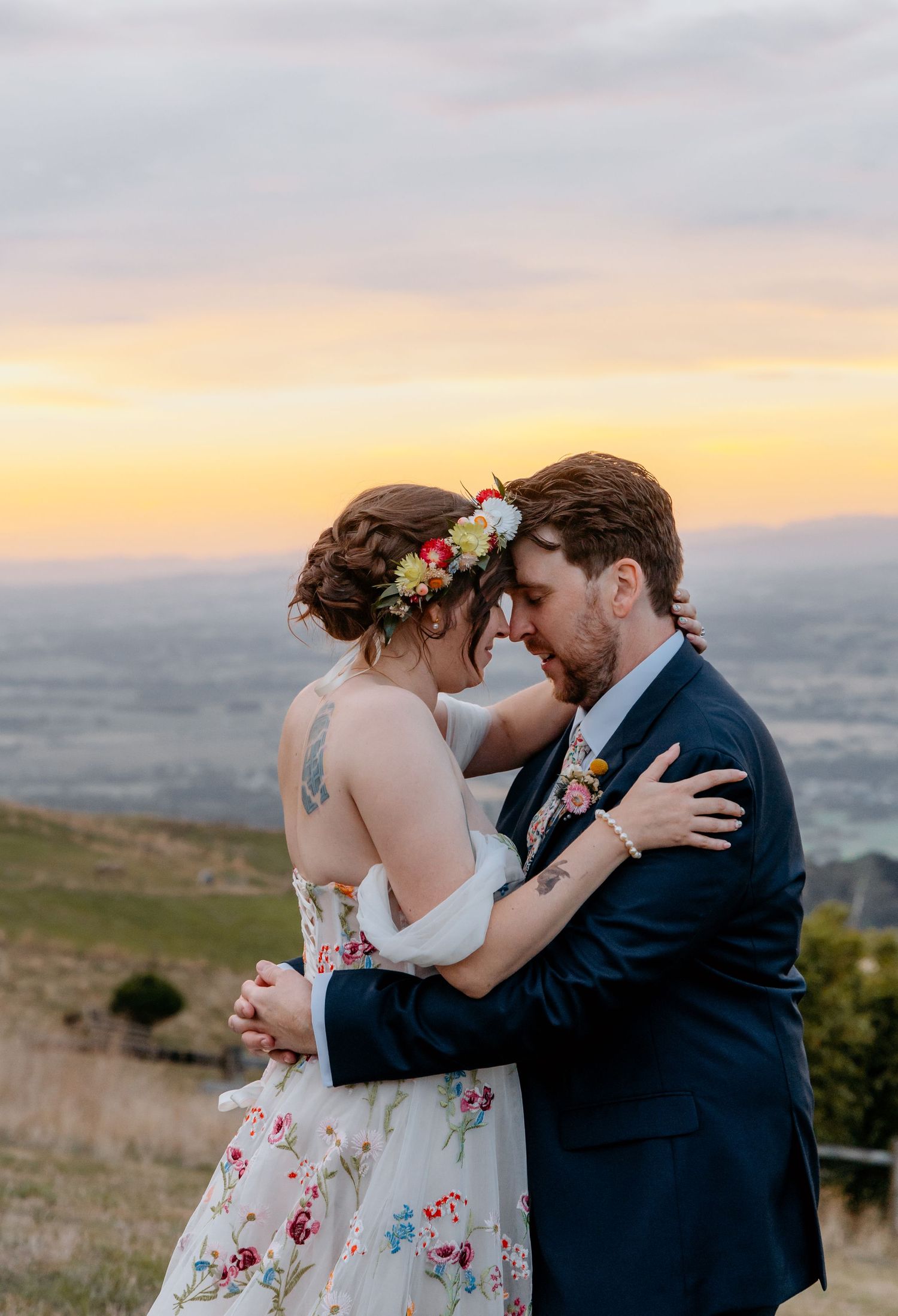 A bride and groom share a romantic sunset embrace outdoors, she wearing a floral gown and flower crown.