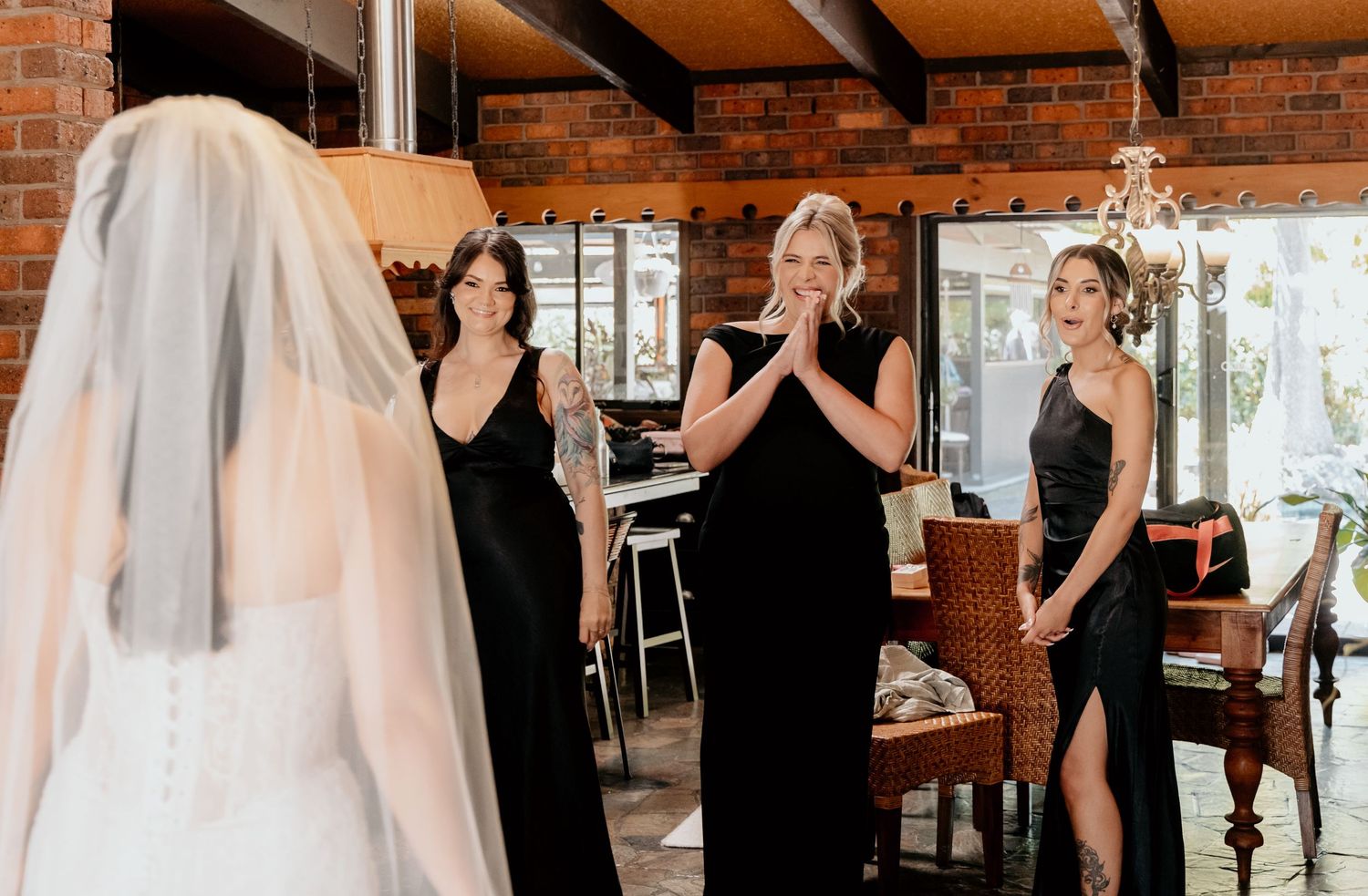 Bridesmaids in black dresses react emotionally as bride in white gown and veil reveals her wedding look indoors.