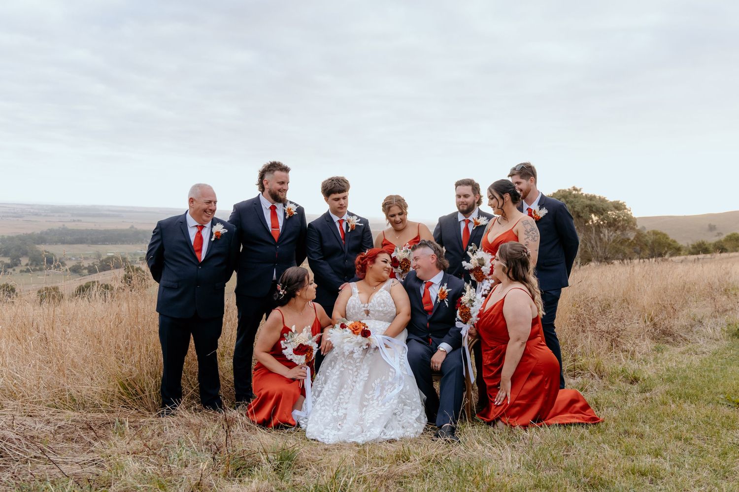 Wedding party in red and navy attire posing in a golden grass field with scenic countryside views.