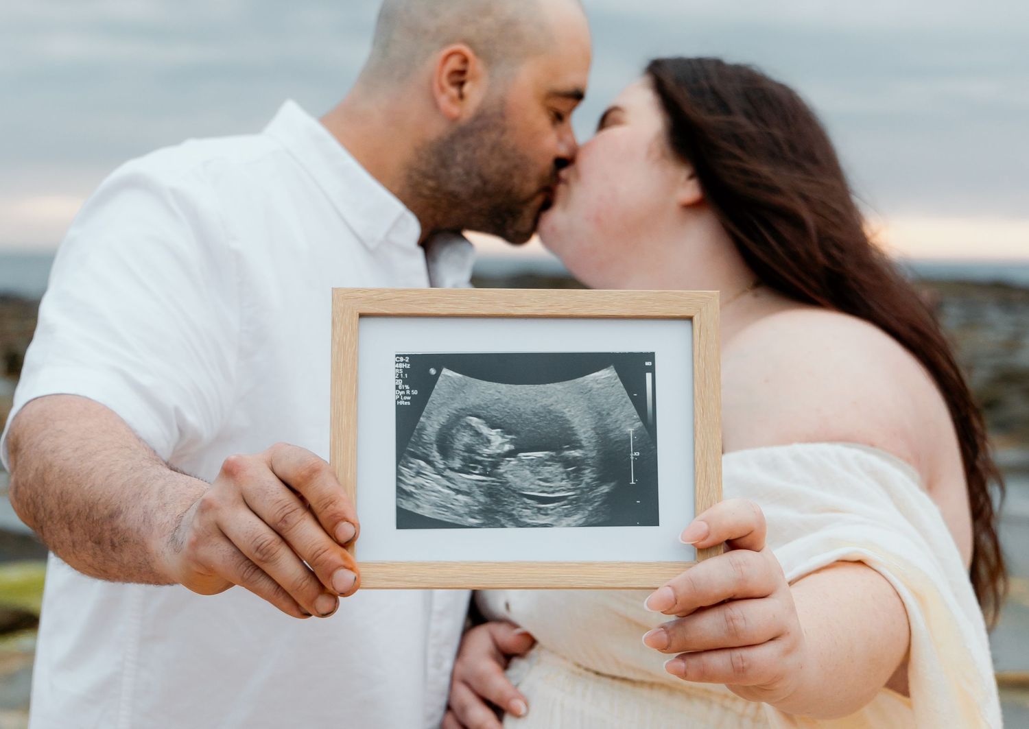 A couple shares a kiss while holding a framed ultrasound photo during a pregnancy announcement photoshoot.