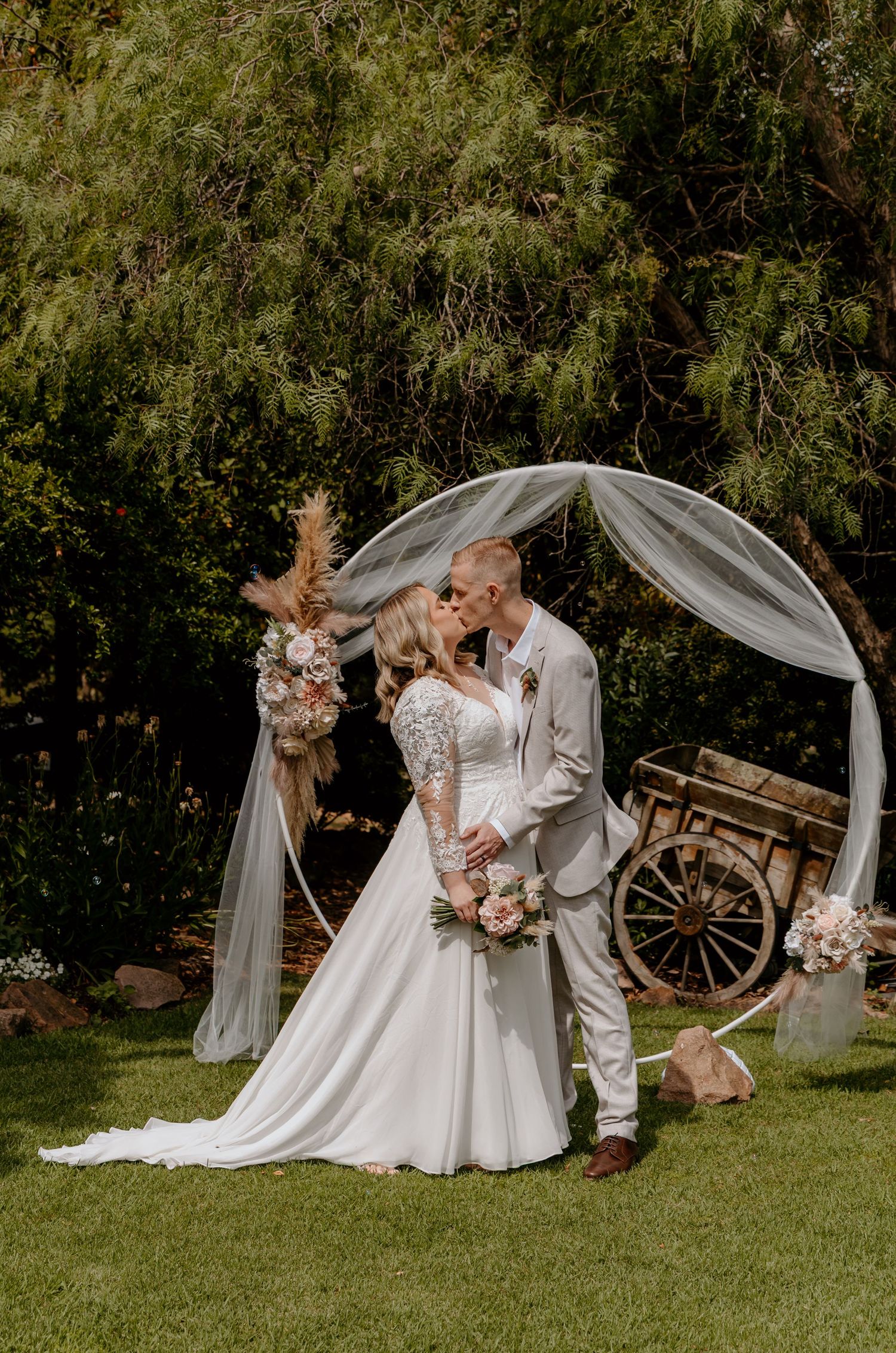 Bride and groom kissing under a circular floral arch with tulle draping in a lush garden wedding ceremony.