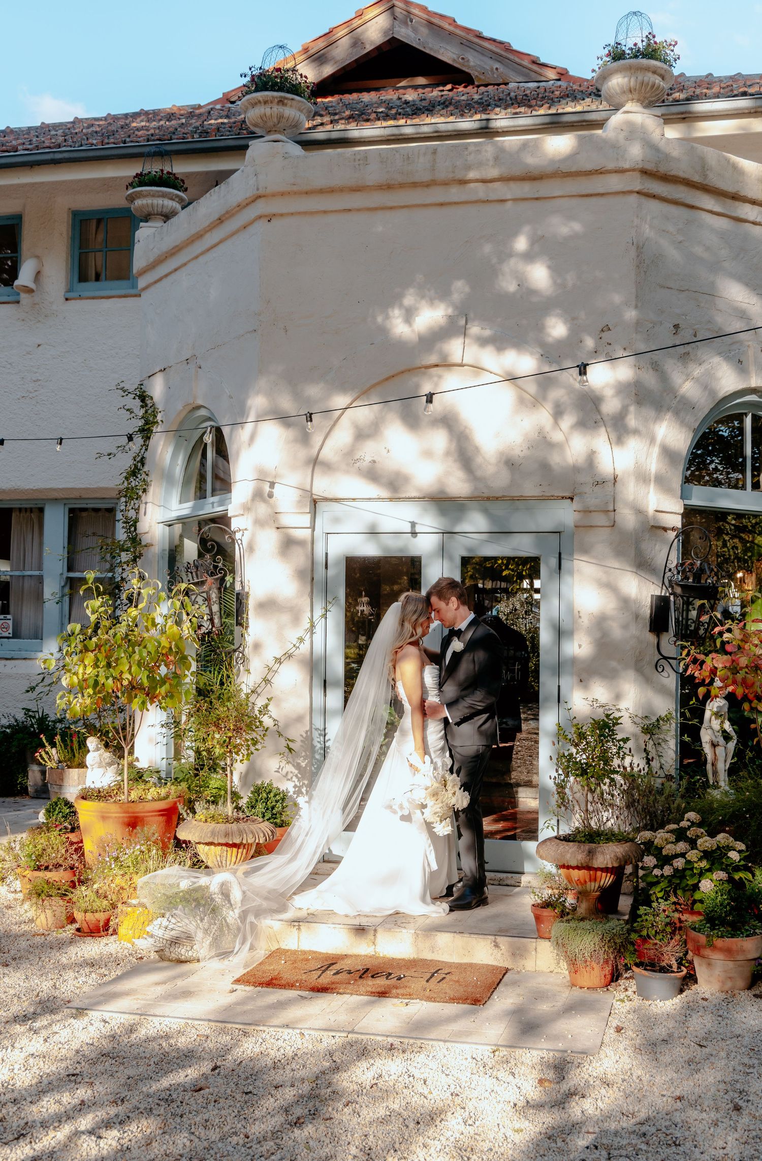 Bride and groom share a romantic moment outside a charming white villa adorned with lush greenery and terracotta pots.