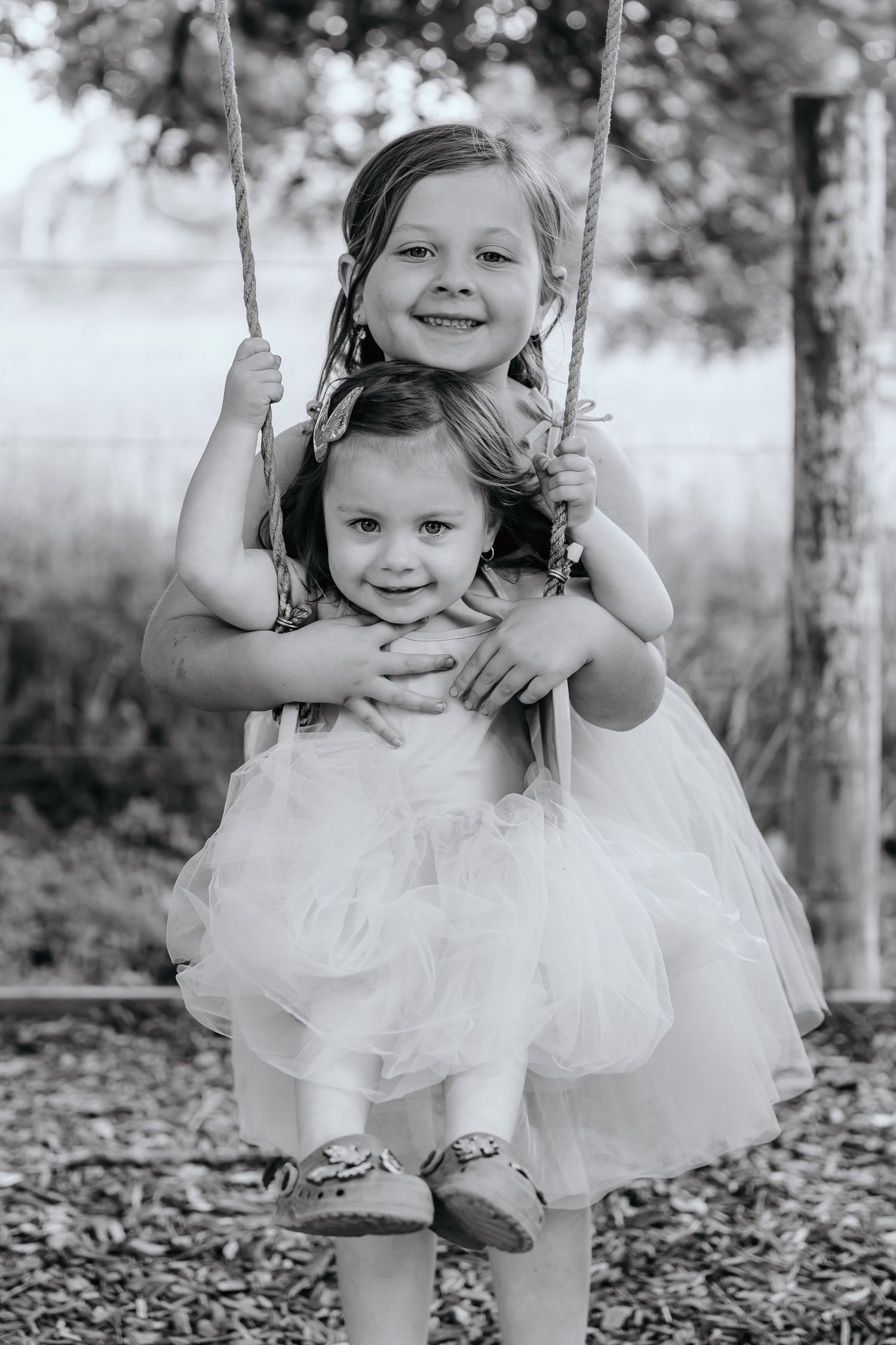 Two young children in white dresses sharing a moment on a wooden swing outdoors.