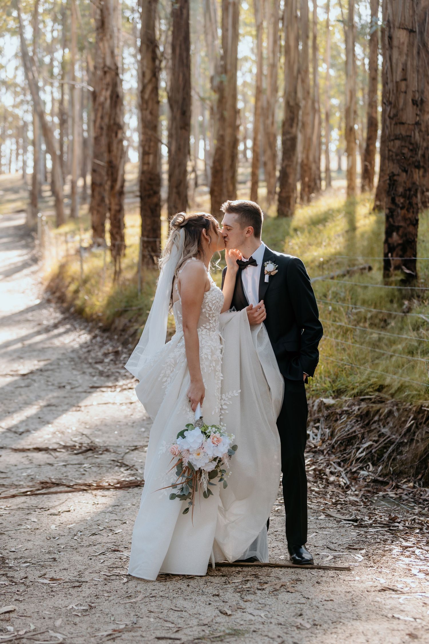 A couple in wedding attire embraces on a sun-dappled forest path while holding a delicate bouquet of flowers.