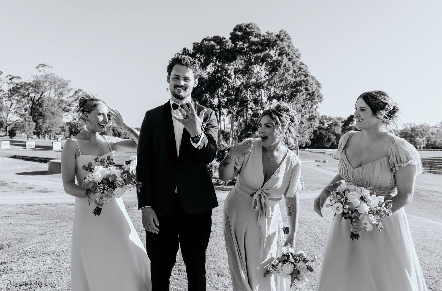 A black and white photo of a wedding ceremony group outdoors laughing together with bouquets and a scenic backdrop.