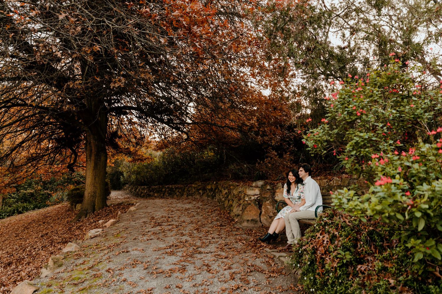 A couple sits on a stone wall along a scenic autumn path surrounded by fall colors.