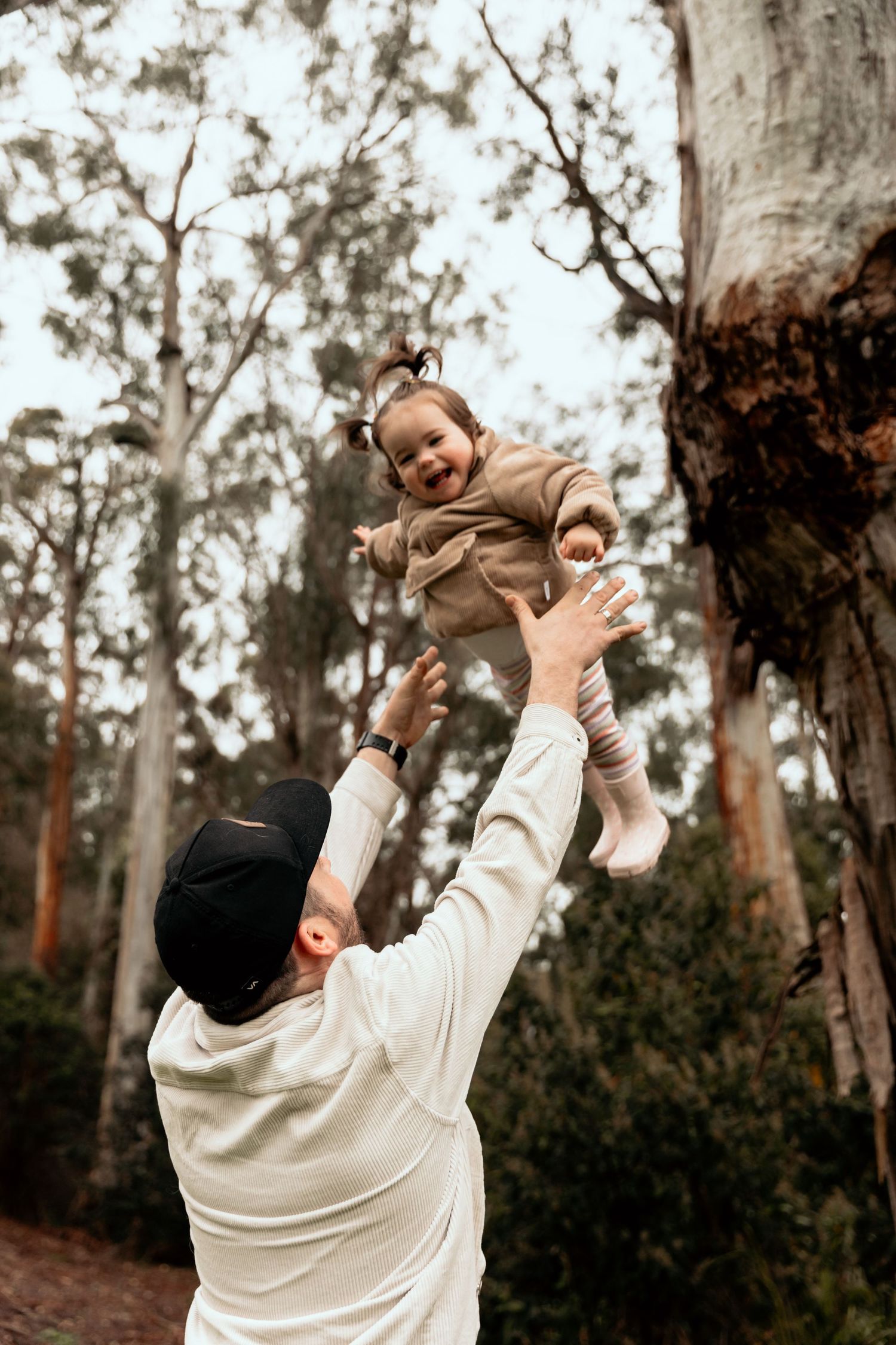 Parent lifting playful toddler in air during outdoor forest adventure.