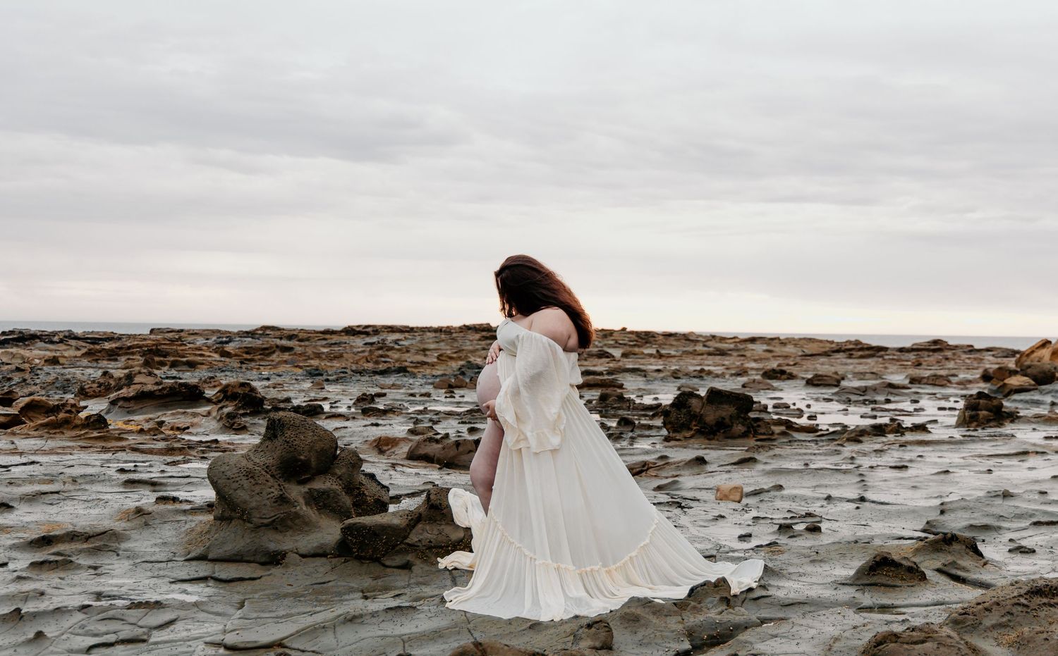 A bride in a flowing white dress walks along a dramatic rocky coastline.