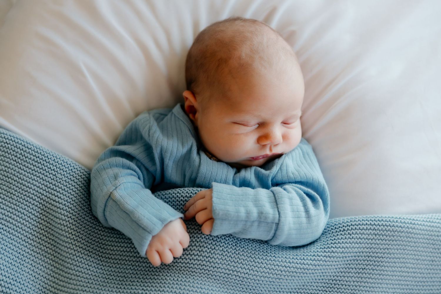 Sleeping newborn in soft blue sweater lying on white and gray bedding.