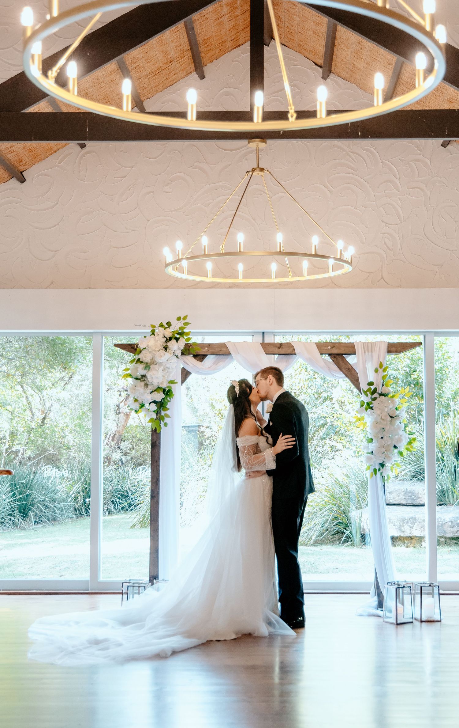 Couple dances in an elegant indoor venue with wooden beams and circular chandelier creating a romantic atmosphere.