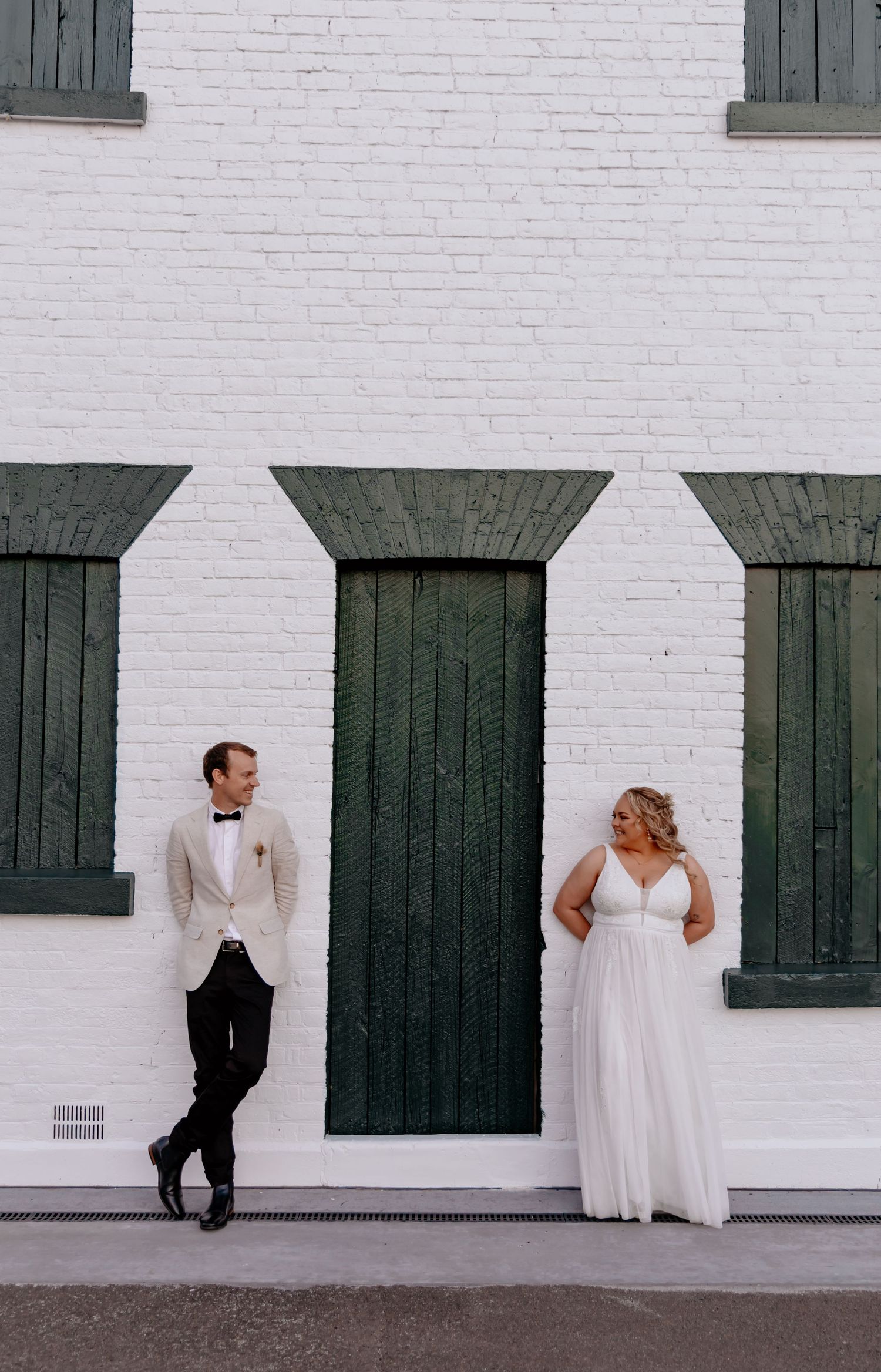 Elegant wedding couple poses against white brick wall with dark green doors creating striking contrast.