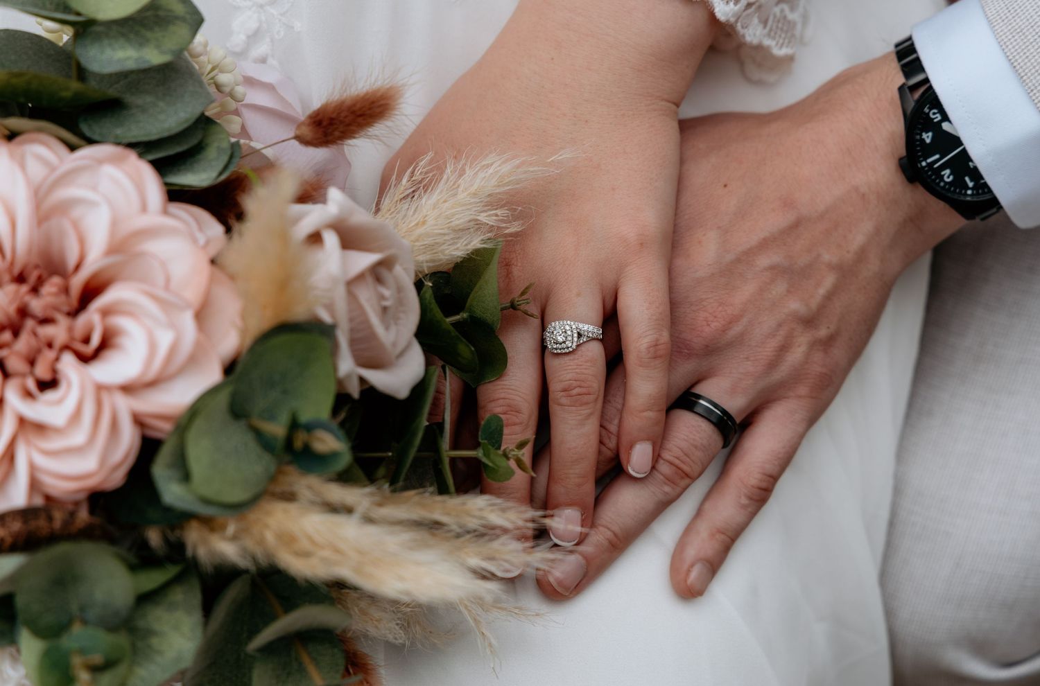 Newlywed couple displaying wedding rings, bride holding pink floral bouquet with eucalyptus greenery.