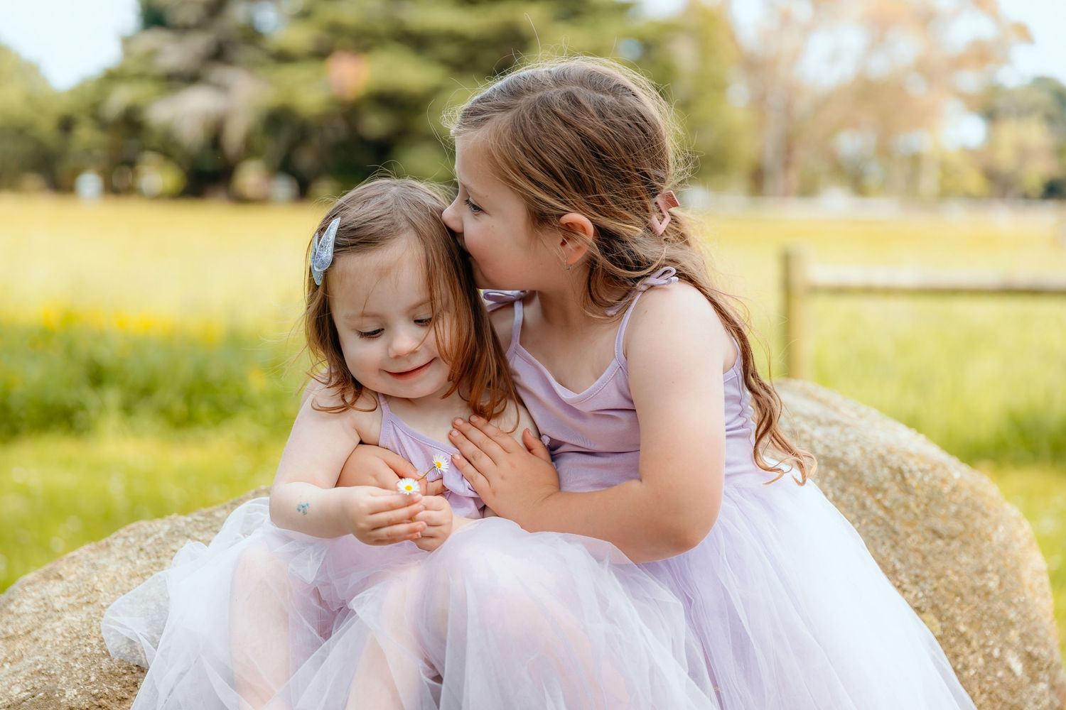 Two children in matching lavender dresses sharing a tender moment on a hay bale.
