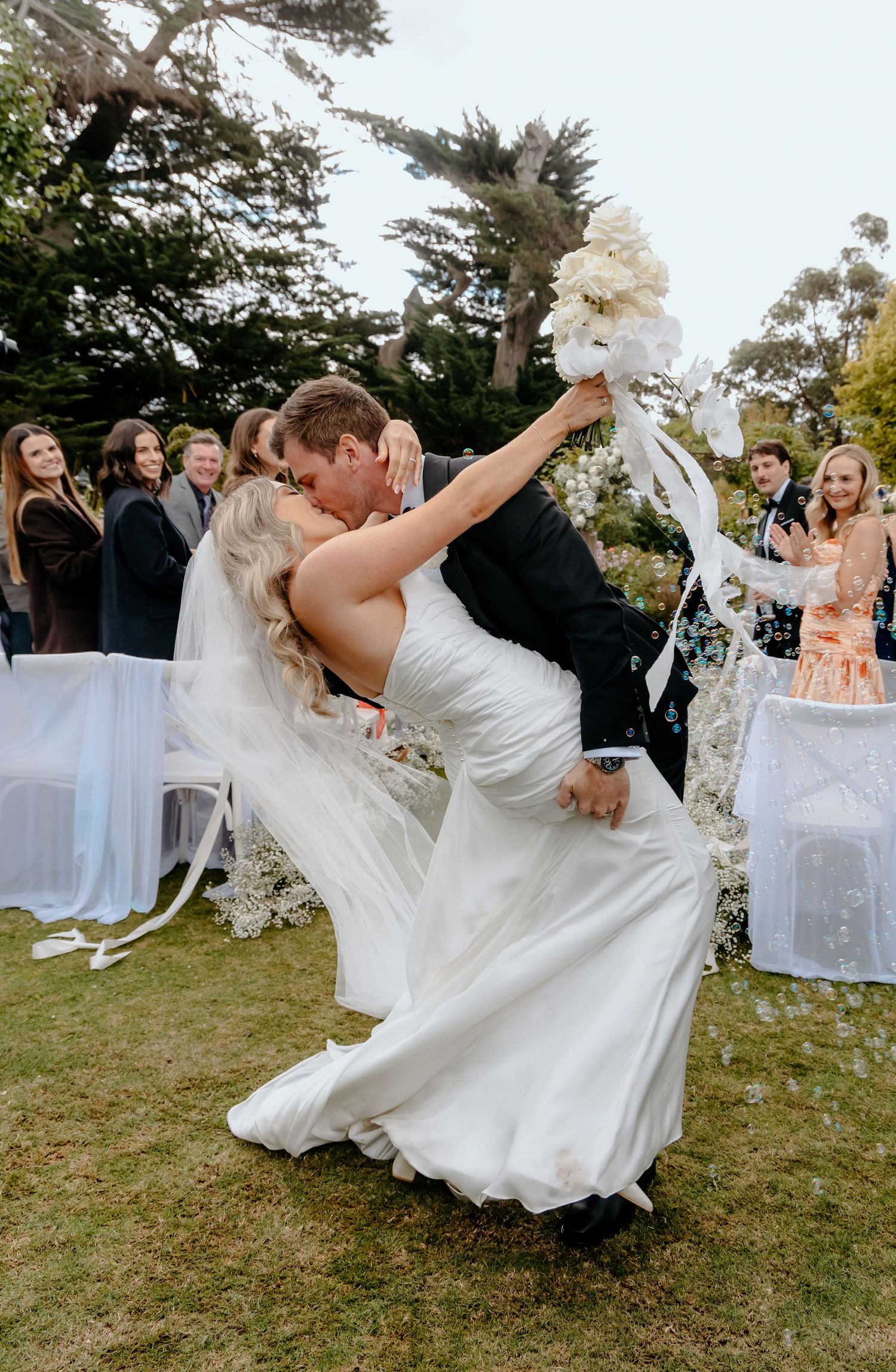 Bride and groom share a romantic dip kiss during outdoor wedding ceremony while guests celebrate.